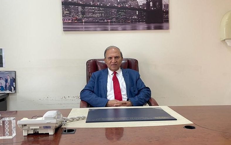 Dr. Naeem Tareen in a suit sits comfortably in a leather chair at an office desk, featuring a phone and a backdrop of a city skyline. about