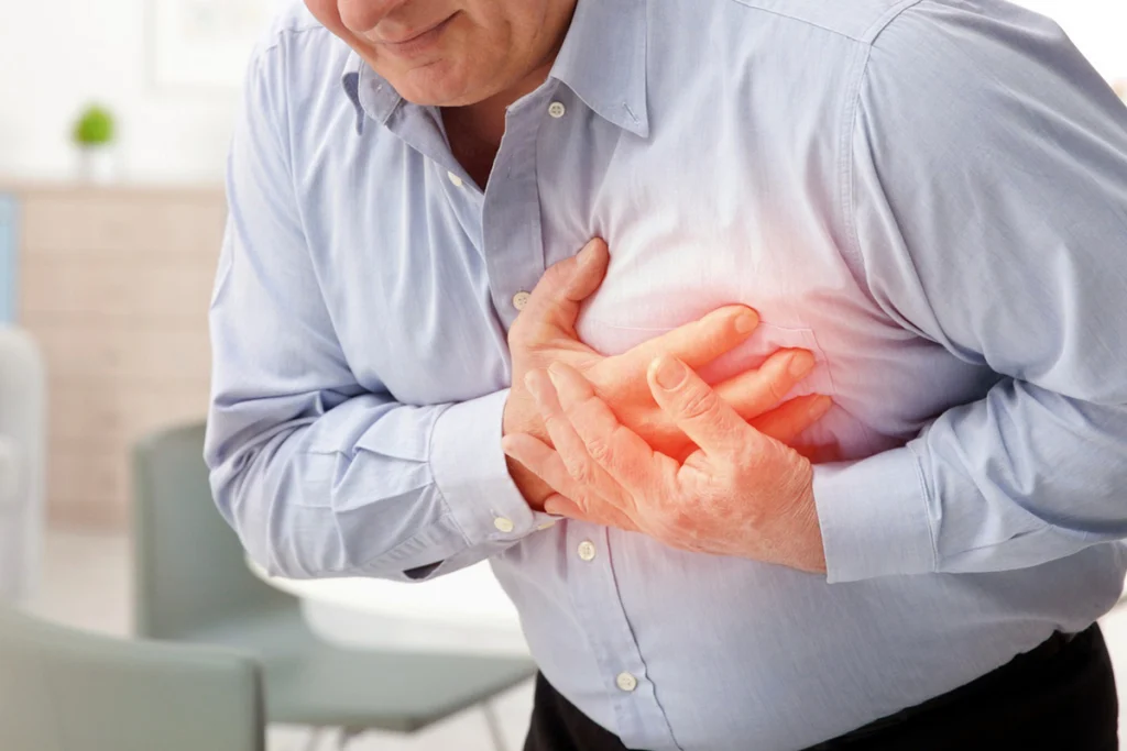 A middle-aged man in a blue shirt grips his chest in pain, suggesting a possible heart issue or discomfort. Bright light highlights the area.
