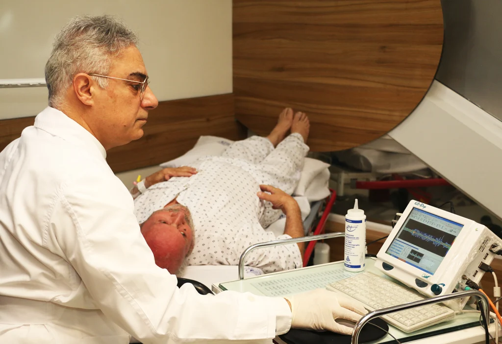 A doctor in a white coat examines a patient lying in a hospital bed, while a medical monitor displays data beside them.