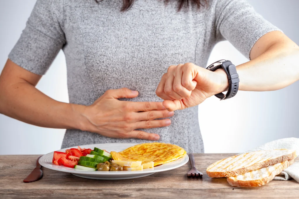 A woman checks her watch while holding her stomach, with a plate of salad, omelette, and bread in front of her.