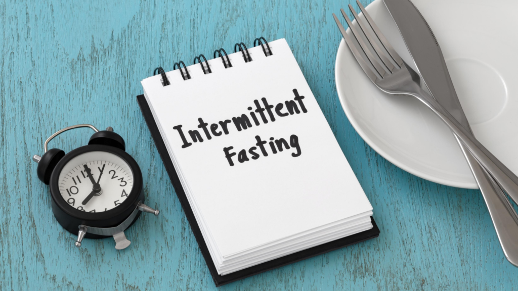 A notepad titled "Intermittent Fasting" beside a black alarm clock and a plate with cutlery on a turquoise wooden table. Intermittent Fasting Benefits for Better Health and Wellness.