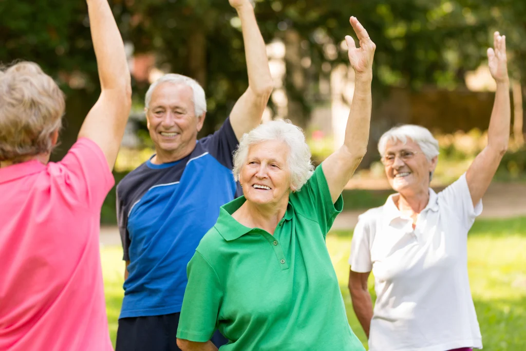 Four elderly individuals are engaged in a group exercise outdoors, raising their arms in unison, surrounded by greenery and trees.