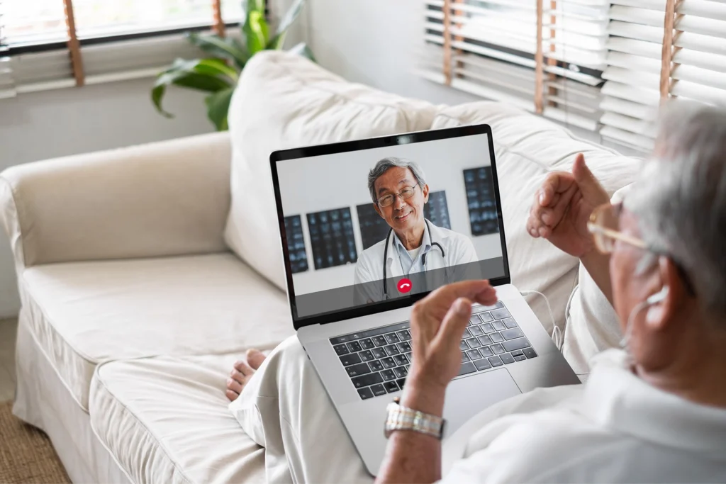 An elderly person engages in a virtual consultation on a laptop, sitting comfortably on a couch in a bright room.