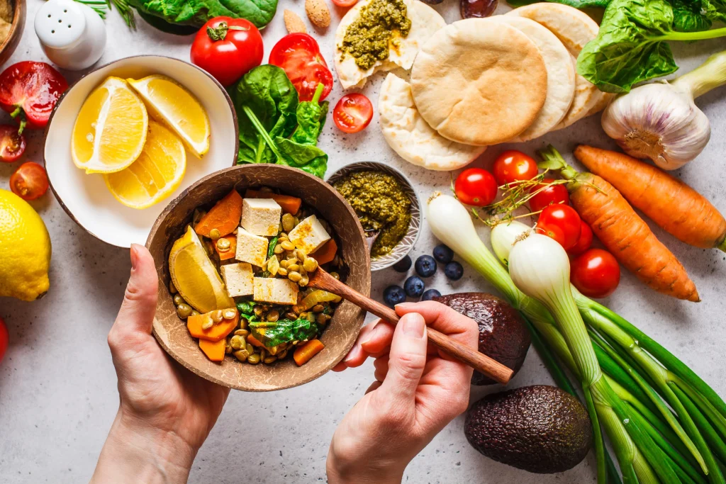 A hand holds a bowl with lentils and tofu, surrounded by fresh vegetables, lemons, and pita, showcasing vibrant, healthy ingredients.