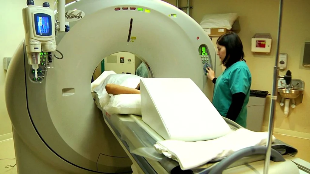 A patient lies on a table in a medical imaging room, preparing for a CT scan, while a technician operates the machine.
