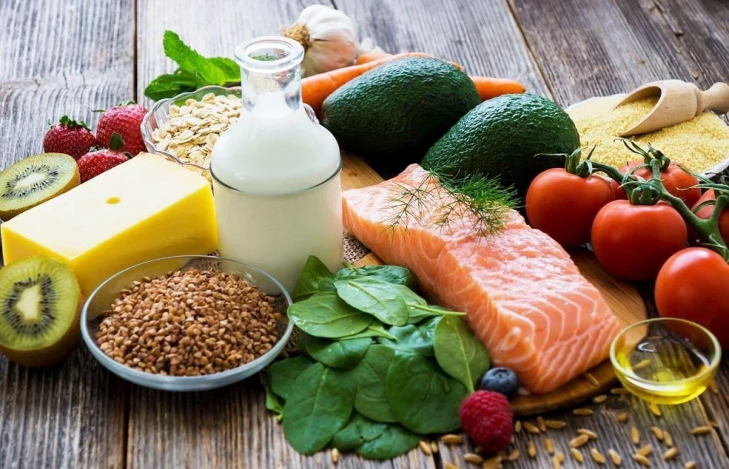 A vibrant assortment of fresh foods including salmon, dairy, vegetables, fruits, and grains arranged on a wooden surface.