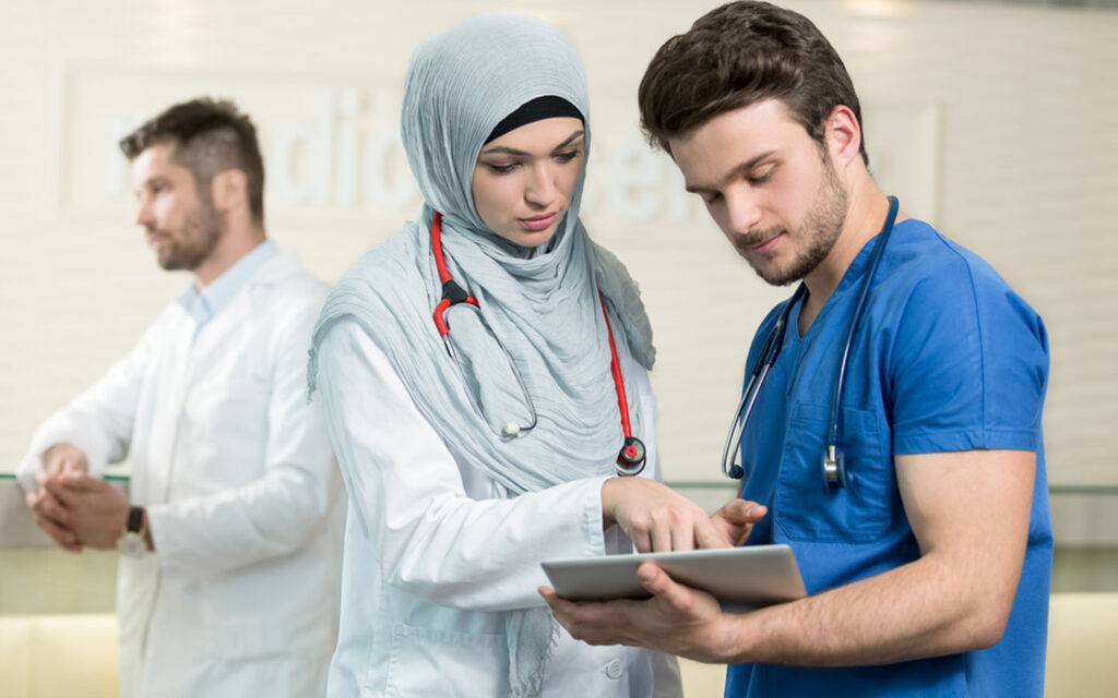 A female healthcare professional in a hijab discusses patient information on a tablet with a male colleague in scrubs.