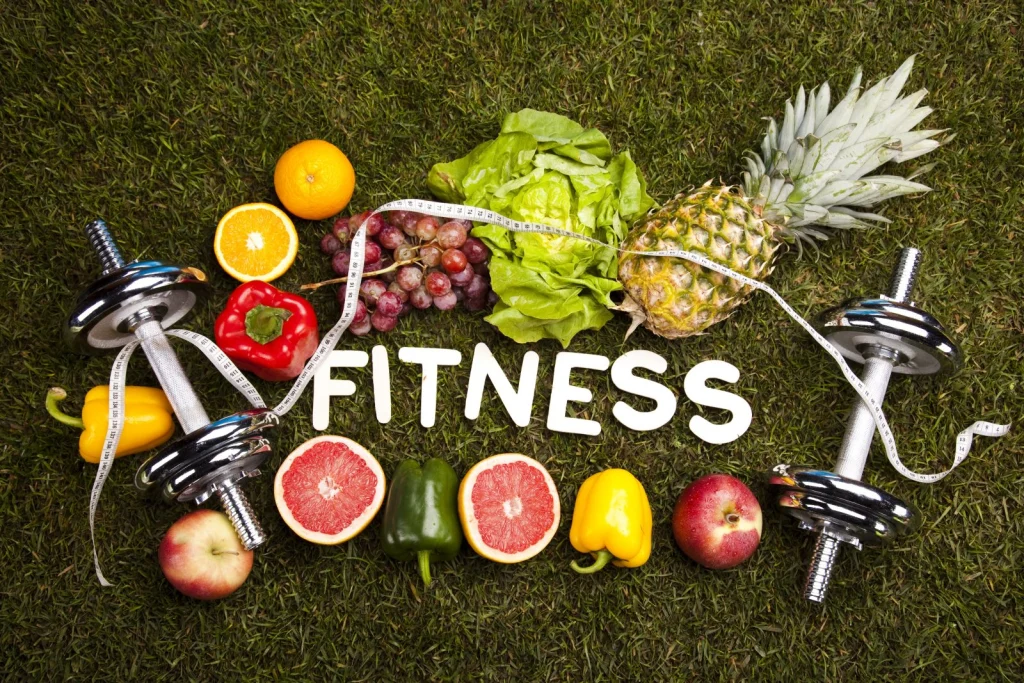 A flat lay of fresh fruits, vegetables, and dumbbells arranged on grass, featuring the word "FITNESS" and a measuring tape.