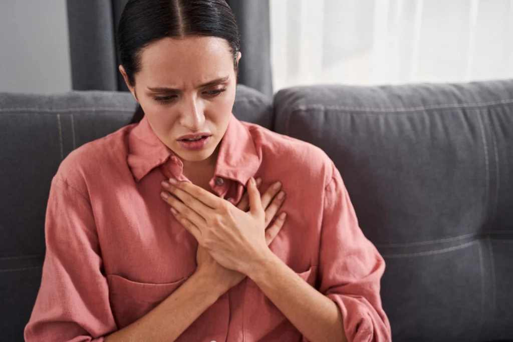 A Lady wearing a pink shirt sits on a grey couch, resting their hands on their chest, conveying a sense of distress or discomfort.